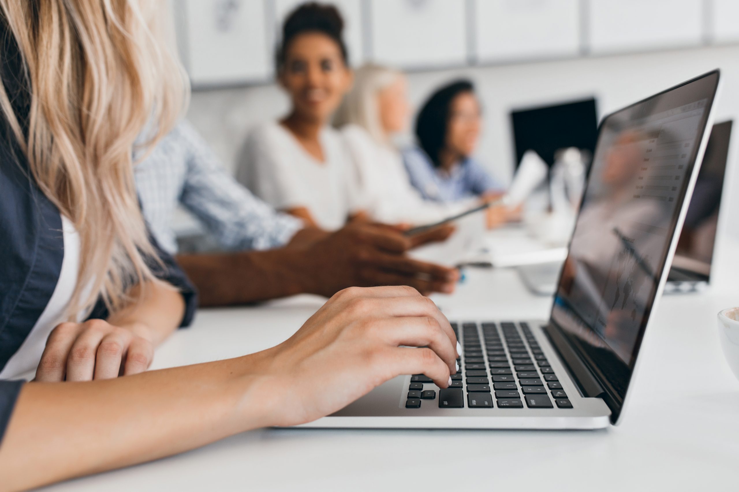 Blonde woman with elegant hairstyle typing text on keyboard in office. Indoor portrait of international employees with secretary using laptop on foreground..